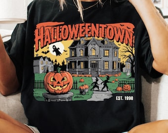 May include: Black t-shirt featuring a Halloween-themed graphic. The design includes a jack-o'-lantern, a haunted house, and the text "Halloweentown" with "EST. 1998". The shirt has a relaxed fit.