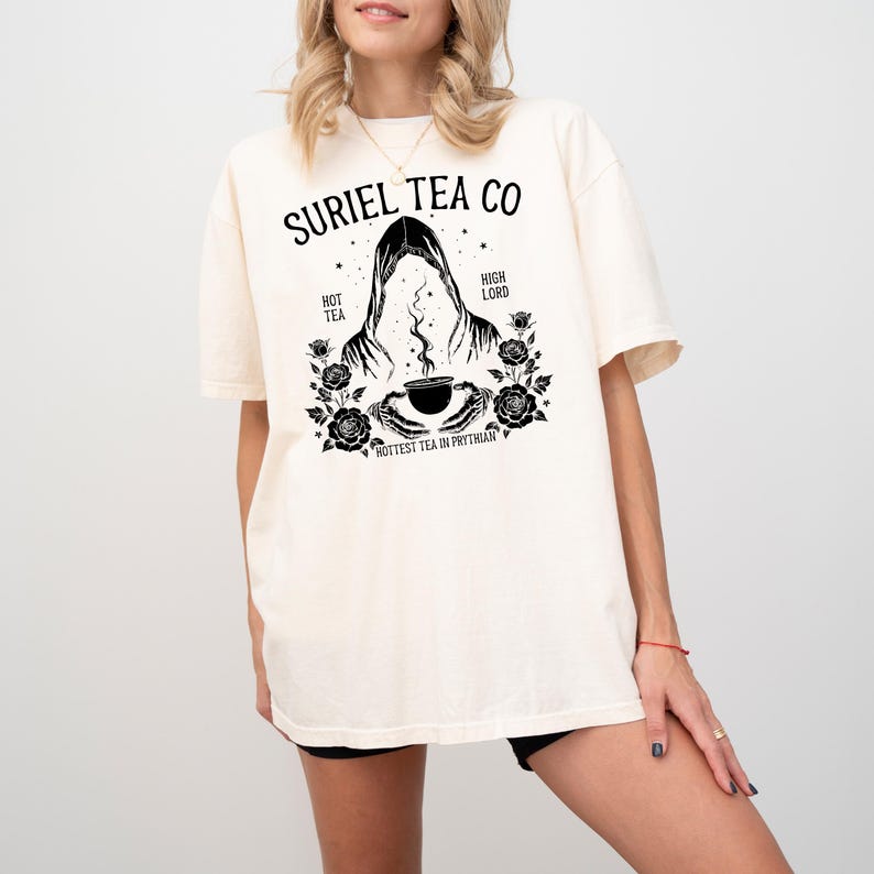 May include: Cream-colored t-shirt featuring a black graphic design. The design includes the text "SURIEL TEA CO" above an illustration of a hooded figure holding a cup of tea, surrounded by roses. Additional text reads "HOT TEA" and "HIGH LORD".