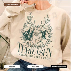 Puede incluir: Una sudadera beige con un diseño gráfico verde de un ciervo geométrico con montañas y estrellas en una corona. El texto "Terrasen Home of the Stag" está debajo del gráfico.