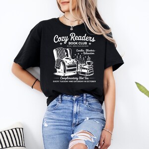 Puede incluir: Camiseta negra con un gr&aacute;fico blanco para un club de lectura. El gr&aacute;fico incluye un sill&oacute;n, velas, libros y una taza de caf&eacute;. El texto en la camiseta dice "Cozy Readers Book Club" y "Complimentary Hot Tea Every Tuesday and Saturday in October."