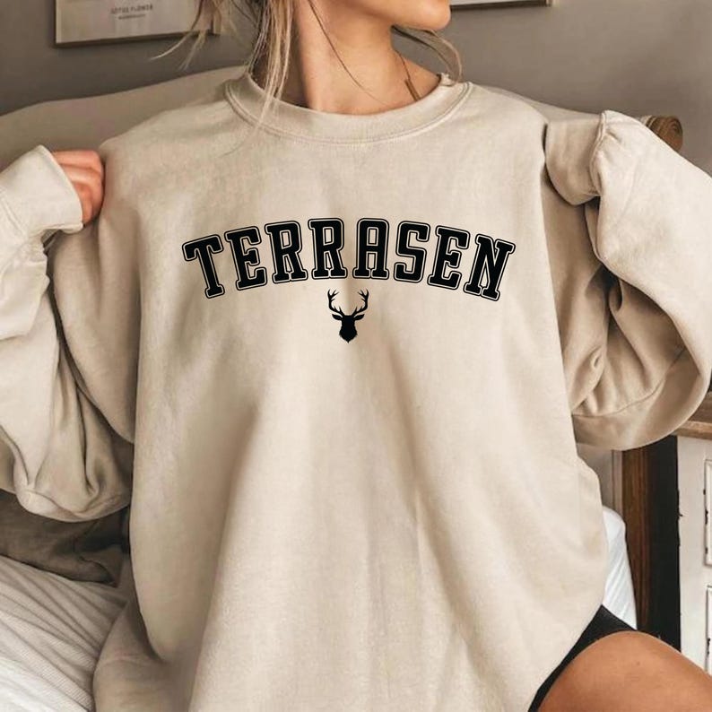 May include: A beige sweatshirt with the text "TERRASEN" in black letters with a black silhouette of a deer with antlers underneath.