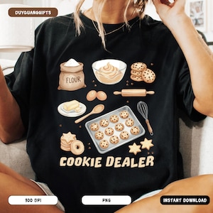 May include: Black t-shirt with a colorful illustration of baking ingredients and cookies. The design includes a flour sack, eggs, butter, a rolling pin, and a baking sheet of cookies. Text on the shirt reads "COOKIE DEALER".
