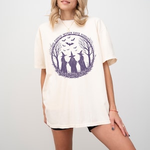 May include: Cream-colored t-shirt featuring a purple graphic of three witches in a forest circle with bats and the text "God Forbid Women Have Hobbies". The shirt has short sleeves and a relaxed fit.