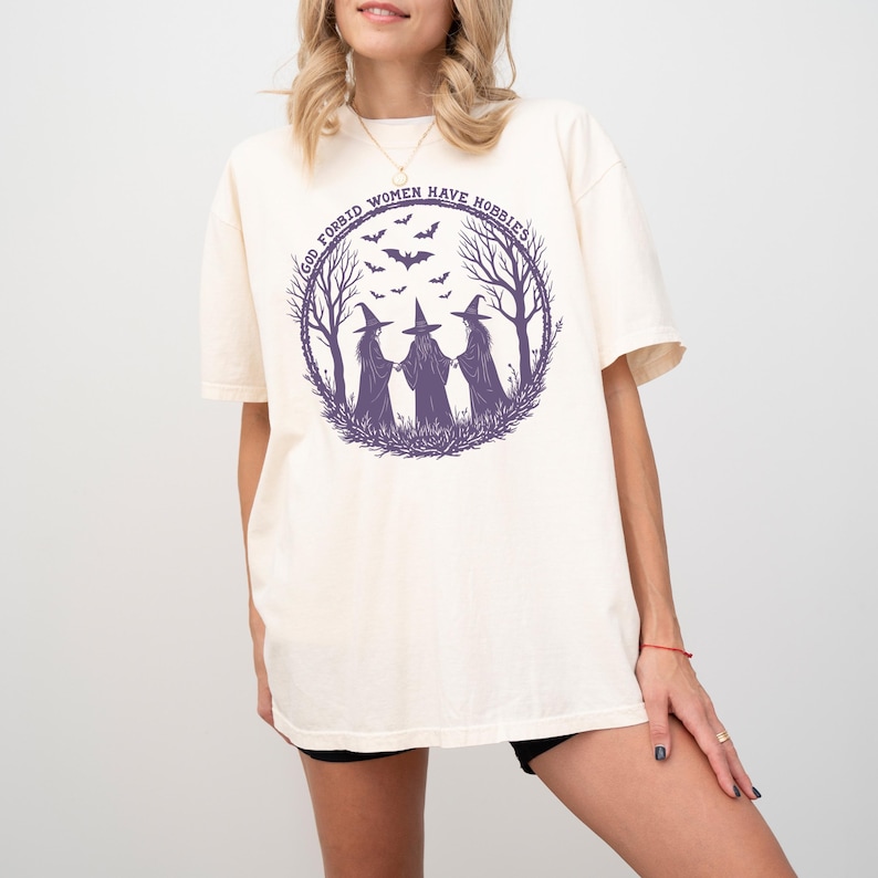 May include: Cream-colored t-shirt featuring a purple graphic of three witches in a forest circle with bats and the text "God Forbid Women Have Hobbies". The shirt has short sleeves and a relaxed fit.