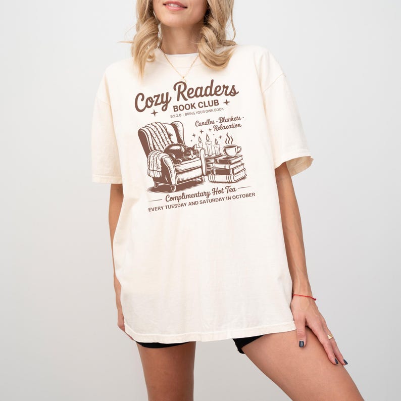 Puede incluir: Camiseta color crema con un gr&aacute;fico marr&oacute;n que representa una escena de lectura acogedora: un sill&oacute;n, libros, velas y una taza de t&eacute;. El texto dice "Cozy Readers Book Club" y "Complimentary Hot Tea Every Tuesday and Saturday in October."