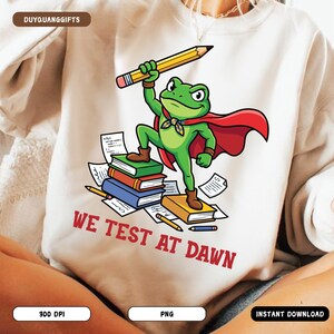 Puede incluir: Sudadera blanca con un dibujo de una rana superhéroe sosteniendo un lápiz. La rana, con una capa roja, está de pie sobre libros con el texto "WE TEST AT DAWN". La rana lleva una pajarita verde.
