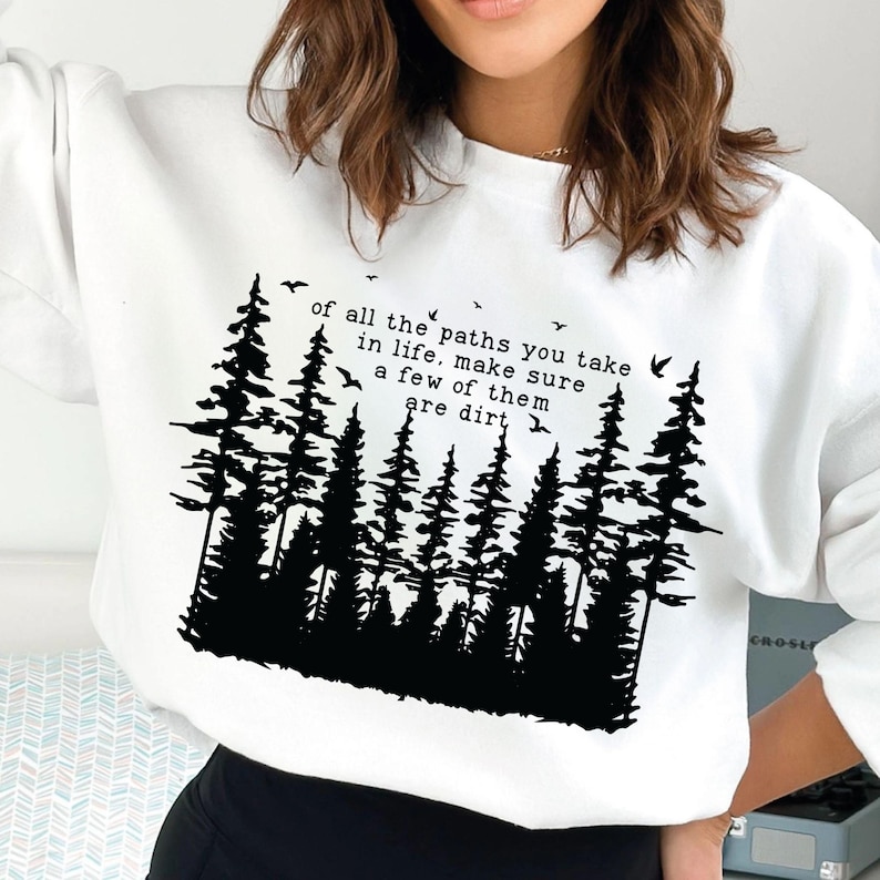 Puede incluir: Sudadera blanca con una silueta negra de un bosque y el texto "of all the paths you take in life, make sure a few of them are dirt".