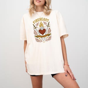 May include: A white t-shirt with a graphic design of a heart on fire with swords crossed behind it. The text "FIREHEART" is above the heart and the text "YOU DO NOT YIELD" is below the heart.