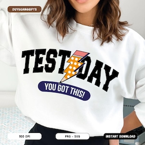 Può includere: Felpa bianca con testo nero che recita "Test Day" con un grafico di fulmine giallo e rosa e il testo "You Got This!" in uno stendardo blu.