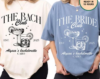 May include: Two white t-shirts with black text and graphics. The left shirt says "The Bach Club" with a cocktail glass and a glass of ice. The right shirt says "The Bride Club" with a cocktail glass and a glass of ice. Both shirts say "Est. 2025" and "Alyssa's Bachelorette Cabo".