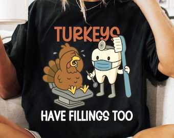 May include: Black t-shirt with a humorous graphic. The design features a cartoon turkey in a dentist's chair and a tooth wearing a mask, holding a toothbrush. Text reads "TURKEYS HAVE FILLINGS TOO."