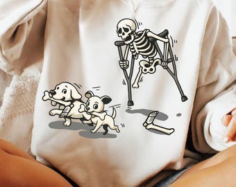 Puede incluir: Sudadera de color crema con un esqueleto de dibujos animados con muletas y dos cachorros juguetones corriendo con huesos. El diseño incluye un hueso desprendido y el texto "DUYQUANGGIFTS".