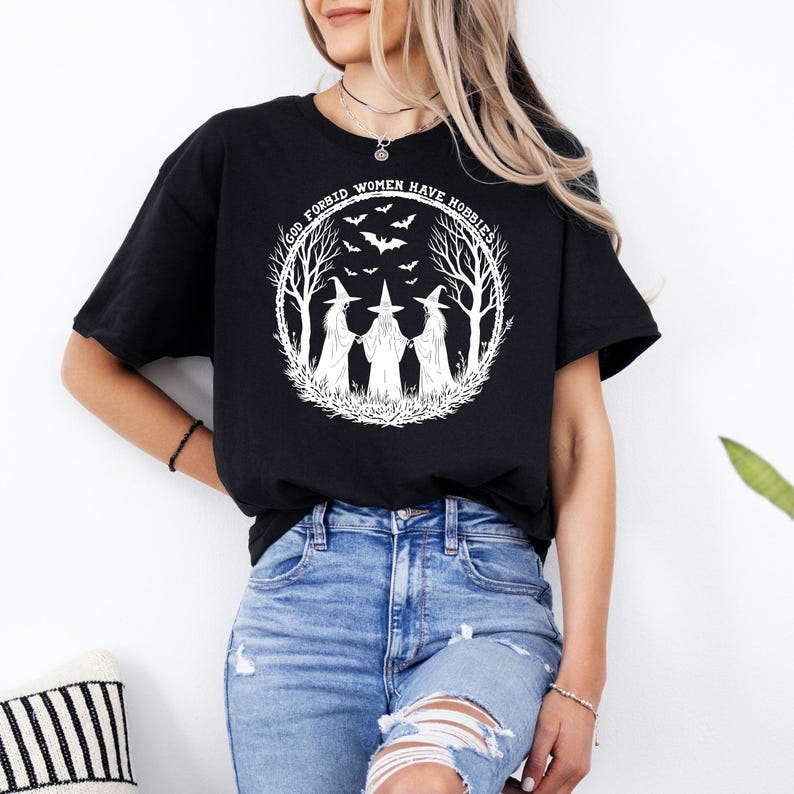 May include: Black t-shirt featuring a white graphic of three witches in a forest circle with bats and the text "GOD FORBID WOMEN HAVE HOBBIES". The design is a whimsical, spooky illustration.
