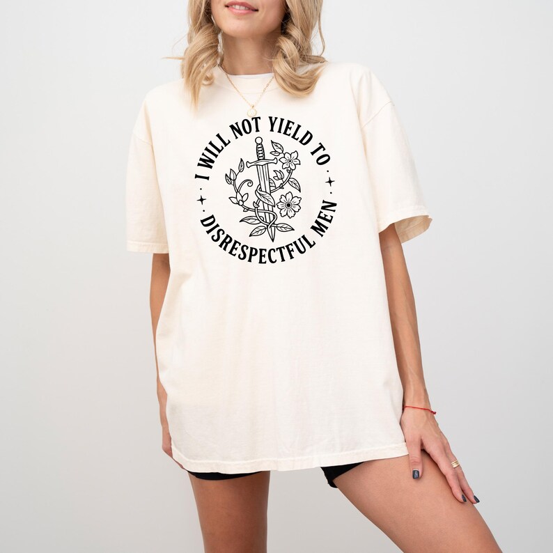 May include: Cream-colored t-shirt featuring a circular graphic with the text "I WILL NOT YIELD TO DISRESPECTFUL MEN." The graphic includes a sword, flowers, and leaves. The shirt has short sleeves and a relaxed fit.