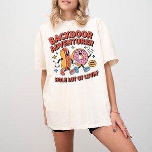May include: Cream-colored t-shirt featuring a cartoon hot dog and donut with the text "BACKDOOR ADVENTURER" and "HOLE LOT OF LOVIN'". The hot dog is orange and the donut is pink with sprinkles. The shirt has a relaxed fit.