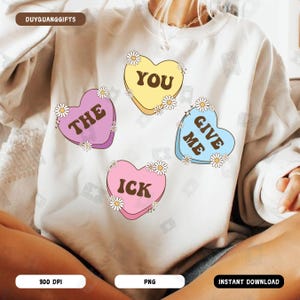 Puede incluir: Una sudadera blanca con cuatro diseños coloridos en forma de corazón. Cada corazón tiene un mensaje diferente: "You", "The", "Give Me" y "Ick".