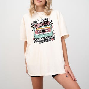 Pu&ograve; includere: Una t-shirt color crema con una grafica a nastro di cassetta in stile retr&ograve;. La grafica dice "Made to Worship" con un cuore rosa e il testo "Let everything that has breath praise the Lord" sotto.