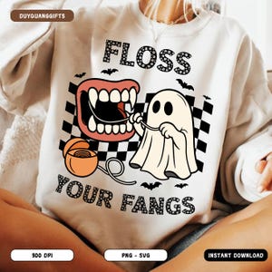 May include: A cream-colored sweatshirt with a Halloween-themed graphic. The design features a cartoon ghost flossing a set of fangs, with the words "FLOSS YOUR FANGS" in a playful font. Black bats and a checkered background complete the design.