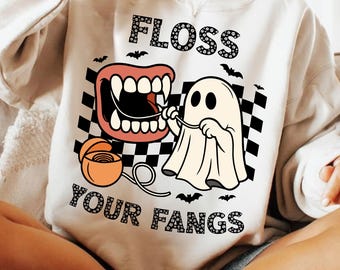 May include: A cream-colored sweatshirt with a Halloween-themed graphic. The design features a cartoon ghost flossing a set of fangs, with the words "FLOSS YOUR FANGS" in a playful font. Black bats and a checkered background complete the design.