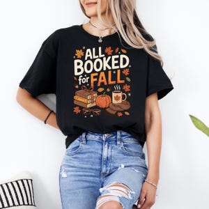 May include: Black t-shirt featuring the text "ALL BOOKED for FALL" in white and orange. The design includes books, a pumpkin, a coffee mug, and autumn leaves. A cosy autumn-themed graphic.