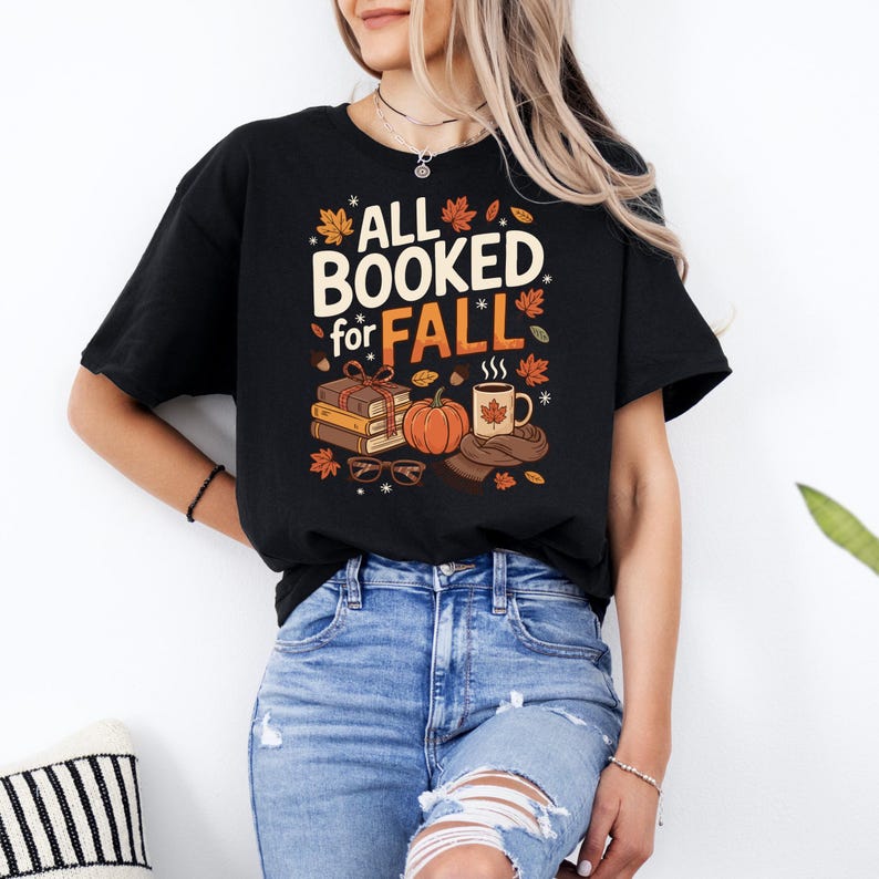 May include: Black t-shirt featuring the text "ALL BOOKED for FALL" in white and orange. The design includes books, a pumpkin, a coffee mug, and autumn leaves. A cosy autumn-themed graphic.