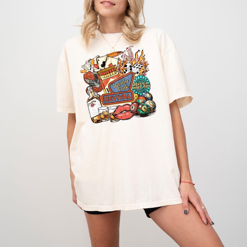 K&ouml;nnte beinhalten: Cremefarbenes T-Shirt mit farbenfroher Grafik. Das Design zeigt den Text "Thely Oak Revival", eine Spirituosenflasche, W&uuml;rfel, einen Mund und Billardkugeln. Das Shirt hat kurze &Auml;rmel und eine lockere Passform.