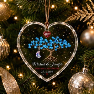 Op de afbeelding: Een hartvormig transparant ornament met een bruine boom met blauwe bladeren, een hangende halve maan bedel en een klein rood hartje met het label 40th. Tekst: Michael & Jennifer, WEDDING ANNIVERSARY en 25.11.1986, opgehangen aan een bruin koord.