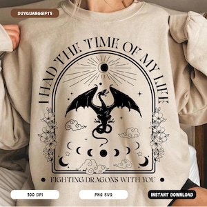 May include: Beige sweatshirt featuring a black dragon design. The graphic includes the text "I HAD THE TIME OF MY LIFE" and "FIGHTING DRAGONS WITH YOU". The design also has moons, clouds, and floral details. A comfortable, casual item of clothing.