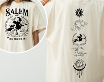 May include: Cream-colored t-shirt with a Salem 1692 witch graphic on the front and a series of celestial and witch-themed illustrations on the back. The front text reads "Salem 1692" and "They Missed One".