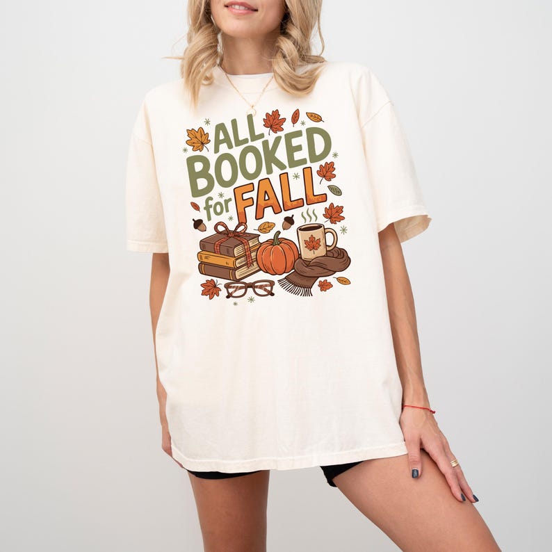May include: A cream-coloured t-shirt featuring a fall-themed graphic. The design includes the text "ALL BOOKED for FALL" alongside illustrations of books, a pumpkin, a coffee mug, and autumn leaves. The shirt is a casual, short-sleeved style.