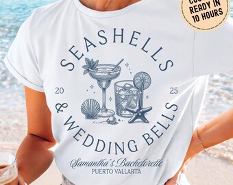 Può includere: T-shirt bianco con un design grafico blu con la scritta "SEASHELLS & WEDDING BELLS". Il design include illustrazioni di un cocktail, un bicchiere di bevanda ghiacciata, una conchiglia e una stella marina. È incluso anche il testo "Samantha's Bachelorette PUERTO VALLARTA".