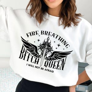 Pode incluir: Camisola branca com um design gr&aacute;fico preto que apresenta uma espada com asas e o texto "Fire Breathing Bitch Queen I Will Not Be Afraid".