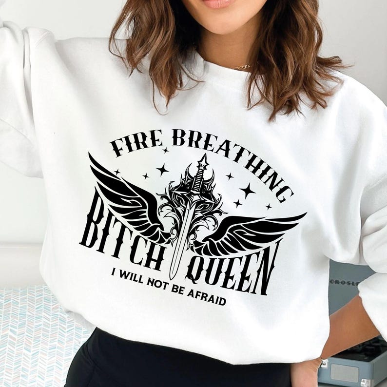 Pode incluir: Camisola branca com um design gr&aacute;fico preto que apresenta uma espada com asas e o texto "Fire Breathing Bitch Queen I Will Not Be Afraid".