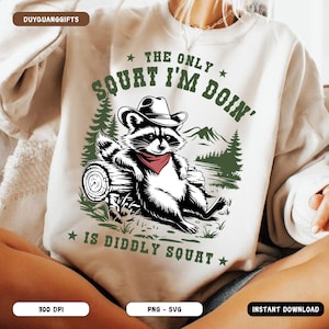 May include: A white sweatshirt with a green, black, and red illustration of a raccoon wearing a cowboy hat. The text "The only squat I'm doin' is diddly squat" is printed on the sweatshirt.