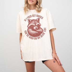 May include: A cream-coloured t-shirt with a raccoon graphic and the text "If I ever get rabies, you're high on my bitin' list". The raccoon illustration has a stern expression and is in a circular design.