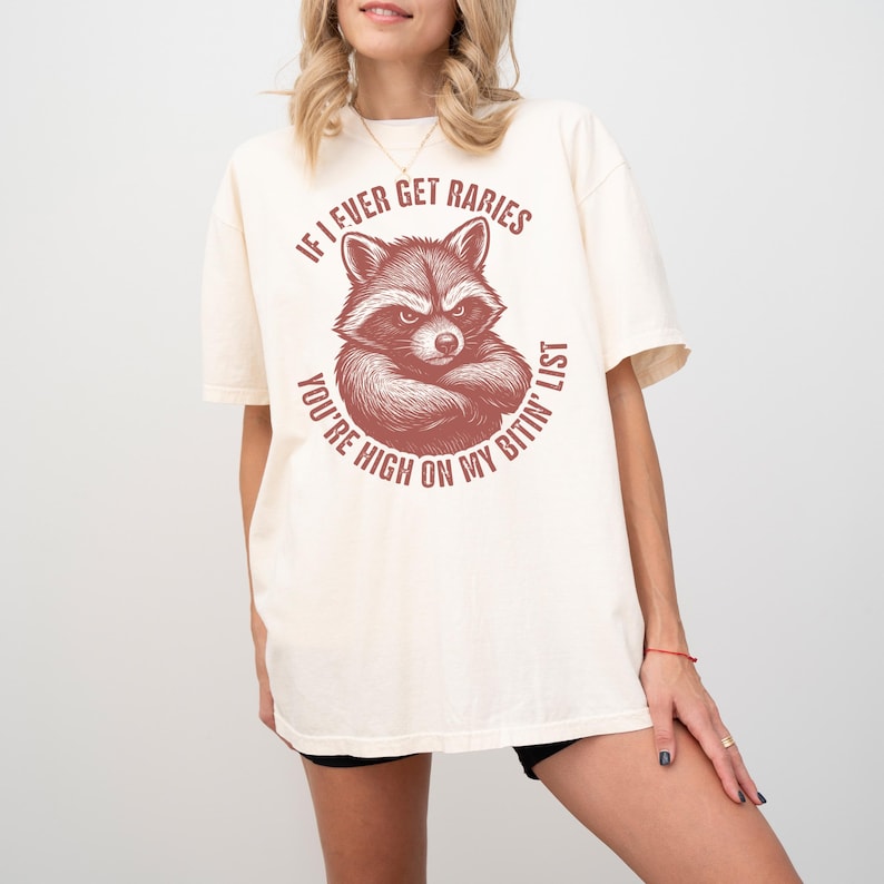 May include: A cream-coloured t-shirt with a raccoon graphic and the text "If I ever get rabies, you're high on my bitin' list". The raccoon illustration has a stern expression and is in a circular design.