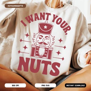 May include: A cream-coloured sweatshirt featuring a burgundy nutcracker graphic and the text "I WANT YOUR. NUTS." The design includes stars and a decorative border. A casual, festive garment.