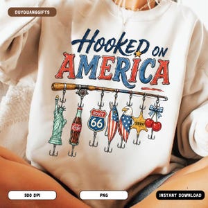 May include: White sweatshirt with the text "Hooked on America" in blue and red. The design features a fishing rod with various patriotic-themed lures, including the Statue of Liberty, a Coca-Cola bottle, and a Route 66 sign.