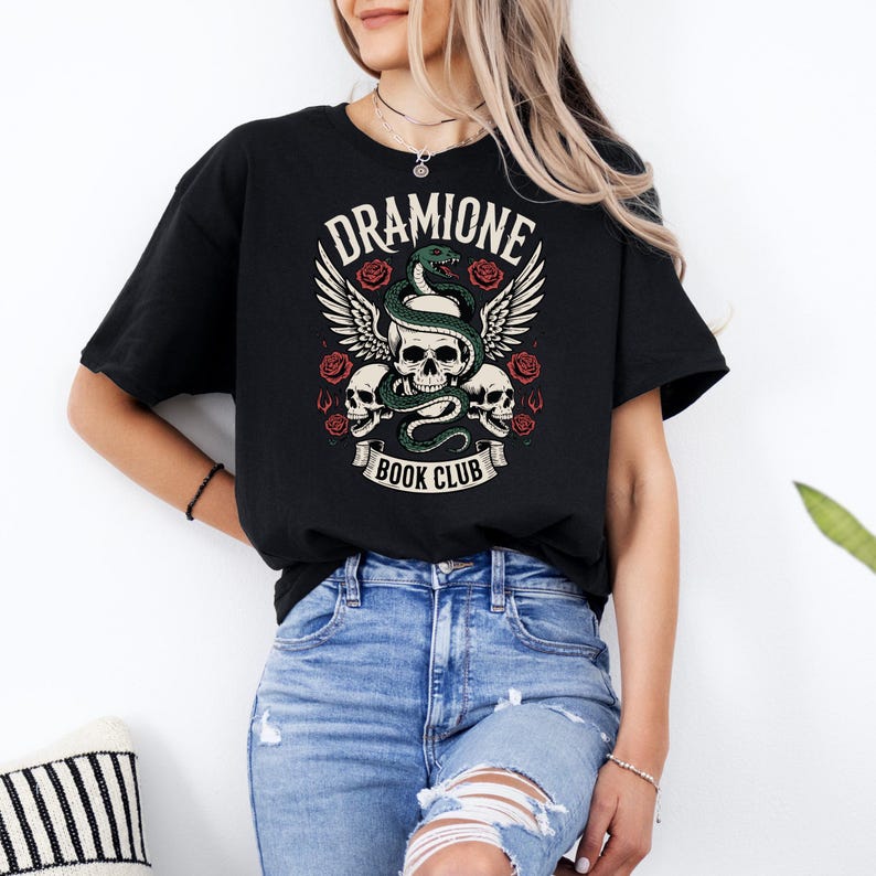 May include: Black t-shirt with a graphic design featuring the text "Dramione Book Club." The design includes a skull, a green snake, wings, and roses. The shirt is made of a soft material and is suitable for casual wear.