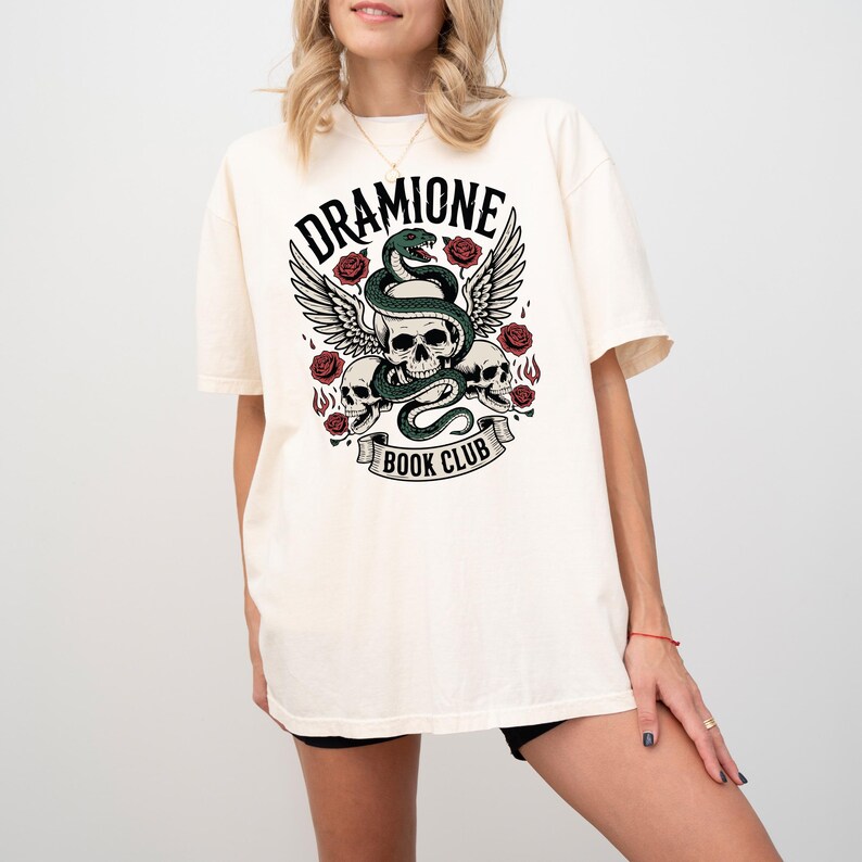May include: Cream-colored t-shirt featuring a graphic design with the text "DRAMIONE BOOK CLUB." The design includes a snake, skulls, roses, and wings. The shirt has short sleeves and a relaxed fit.