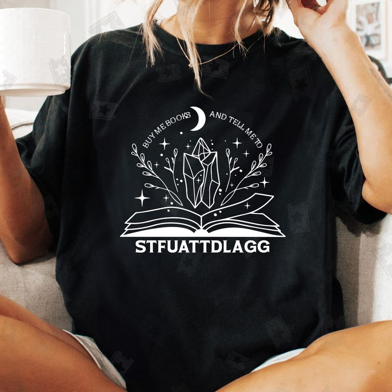 May include: Black t-shirt with a white graphic design of an open book with a crystal and a crescent moon. The text "Buy me books and tell me to STFUATTDLAGG" is printed below the graphic.