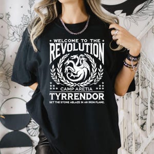 Peut inclure: T-shirt noir avec un motif blanc repr&eacute;sentant un dragon &agrave; trois t&ecirc;tes, une couronne de laurier et le texte "Welcome to the Revolution", "Camp Aretia", "Tyrendor" et "Set the stone ablaze in an iron flame."