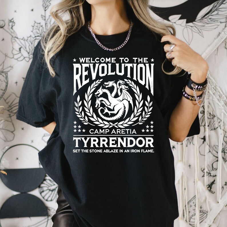 Peut inclure: T-shirt noir avec un motif blanc repr&eacute;sentant un dragon &agrave; trois t&ecirc;tes, une couronne de laurier et le texte "Welcome to the Revolution", "Camp Aretia", "Tyrendor" et "Set the stone ablaze in an iron flame."