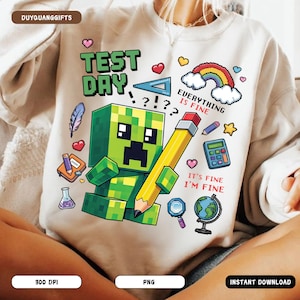 May include: A white sweatshirt featuring a green Minecraft character holding a yellow pencil. The text "Test Day!?!? Everything is Fine" is printed on the sweatshirt. Other school-related graphics on the sweatshirt include a rainbow, a ruler, a calculator, a globe, and a magnifying glass.