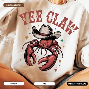 Può includere: Una felpa color crema con un'illustrazione a cartoni animati di un'aragosta rossa con un cappello da cowboy. Il testo "YEE CLAW" è stampato sopra l'aragosta in un font in stile western. Sono visibili anche stelle decorative e il testo "300 DPI", "PNG" e "INSTANT DOWNLOAD".