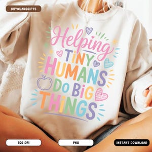 May include: Cream-coloured sweatshirt featuring the phrase "Helping Tiny Humans Do Big Things" in a vibrant, multi-coloured font. The design incorporates hearts, an apple, and sunbursts, suitable for teachers or educators.