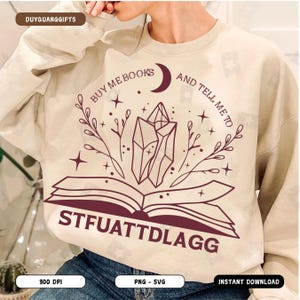 May include: A beige sweatshirt with a brown graphic design of an open book with crystals and stars. The text "BUY ME BOOKS AND TELL ME TO STFUATTDLAGG" is printed on the sweatshirt.
