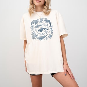 May include: Cream-coloured t-shirt with a circular design of marine animals in shades of blue. The design includes a turtle, dolphin, octopus, whale, and other sea creatures. The shirt has short sleeves and a relaxed fit.