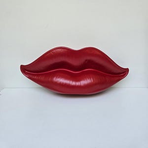 Könnte beinhalten: Ein großes, rotes, glänzendes, geformtes Lippenpaar. Die Lippen sind geschlossen und haben eine glatte, glänzende Oberfläche.