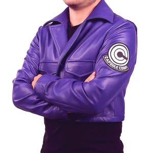 May include: A purple leather jacket with a collared neckline and two front pockets. The jacket features a Capsule Corp patch on the upper left sleeve. The person is wearing a black shirt underneath.